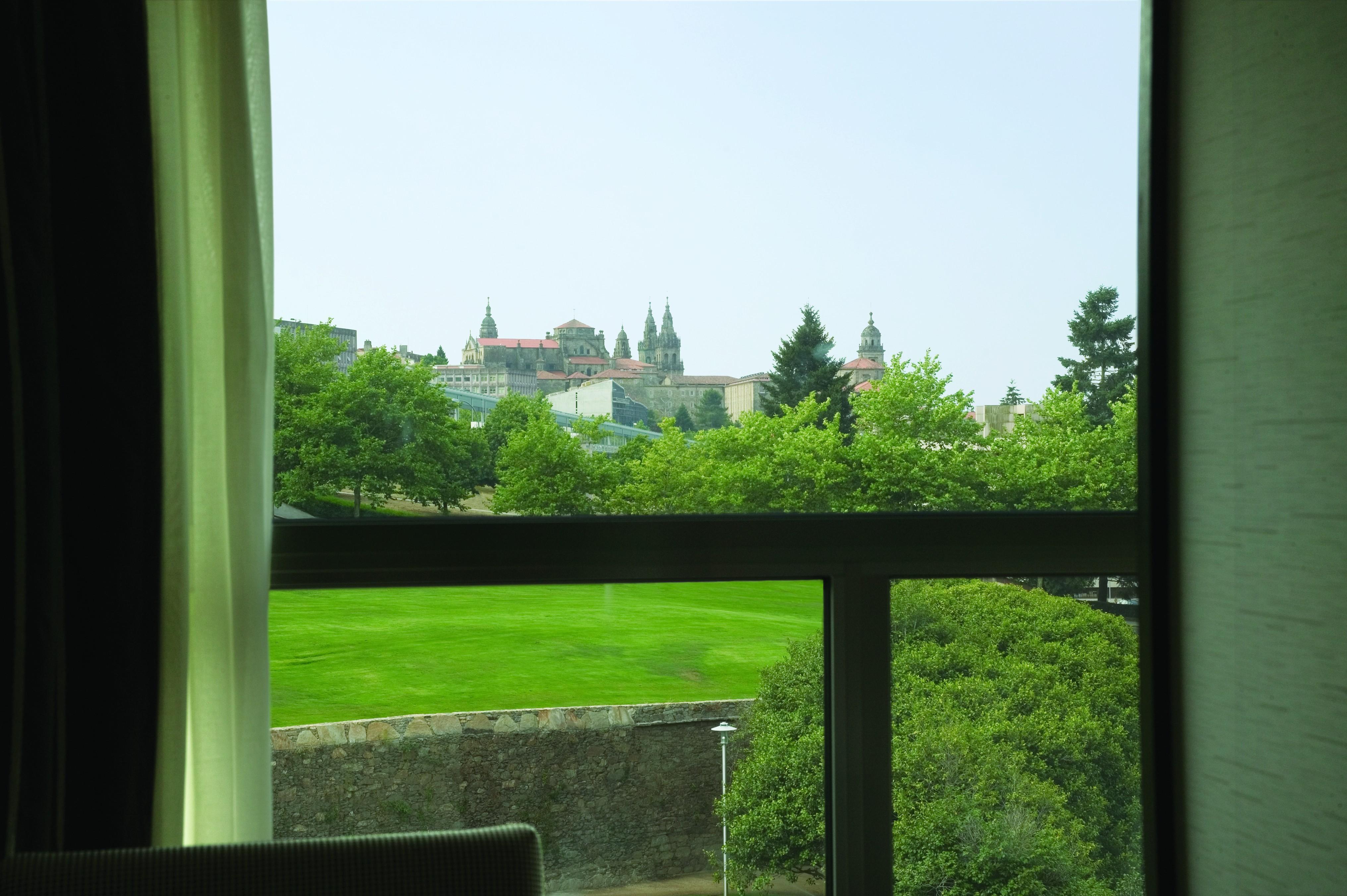 Nh Collection Santiago De Compostela Hotel Exterior photo The photo shows a view from a window, framing a landscape that includes a green area with grass and trees. In the background, there are buildings with distinct architectural features, possibly a historic site or a castle, with towers and spires. The 