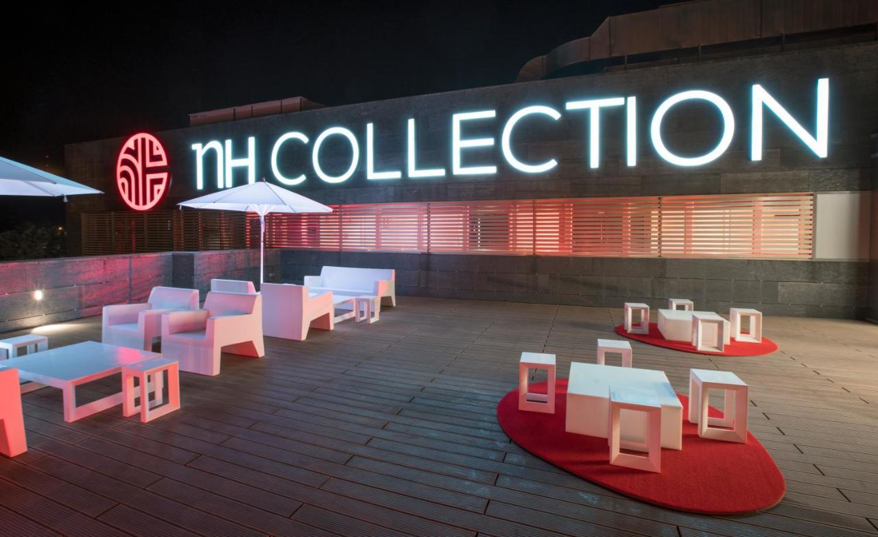 Nh Collection Santiago De Compostela Hotel Exterior photo The photo depicts a rooftop setting featuring modern, minimalist furniture. There are several white lounge chairs and small tables arranged on a wooden deck. A large umbrella stands in the area, providing shade. The backdrop features an illuminated s
